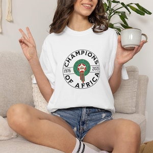 May include: White t-shirt with a circular graphic that reads "Champions of Africa" in black text. The graphic includes a green emblem with red and white accents, and the years 1976 and 2025. The shirt is paired with denim shorts.
