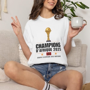 以下が含まれることがあります： 金色のトロフィーと「CHAMPIONS D'AFRIQUE 2025」の文字がプリントされた白いTシャツ。テキストの下には2つの星と旗があります。「COUPE D'AFRIQUE DES NATIONS」の文字も含まれています。