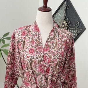May include: A long-sleeved robe featuring a light pink base with a repeating floral pattern in shades of dark pink and olive green. The garment includes a V-neckline, a matching fabric tie belt at the waist, and white piping along the front edges.