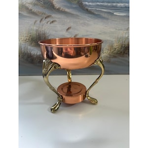 May include: A polished copper bowl with a smooth, reflective surface, supported by three ornate, curved brass legs that feature decorative detailing at the top and connect to a central circular copper base platform for stability.