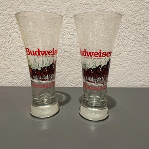 May include: Two clear Budweiser beer glasses. Each glass has a flared shape and a white base. The Budweiser logo is in red, with a graphic of horses and the words "KING OF BEERS" and "CLYDESDALES" in white.