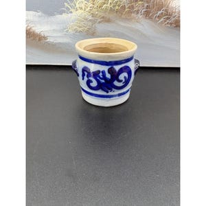 Op de afbeelding: Een kleine, cilindervormige keramische pot met een crèmekleurige basis en een brede, open bovenkant. De buitenkant is voorzien van een handgeschilderd kobaltblauw decoratief patroon van wervelende, abstracte motieven en horizontale banden, met twee kleine handvatten.
