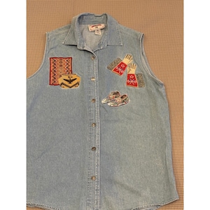 Puede incluir: Una camisa sin mangas de mezclilla azul claro con parches bordados en la parte delantera. Los diseños incluyen una alfombra estampada, una vasija de cerámica con un águila, un par de guantes y un par de zapatos. La camisa tiene cuello clásico.