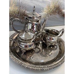 Op de afbeelding: Een verzilverd theeservies bestaande uit een theepot, een suikerpot met deksel en een melkkannetje, gerangschikt op een rond, versierd dienblad. Elk stuk heeft gepolijste metalen oppervlakken, gebogen handvatten en decoratieve voetjes.