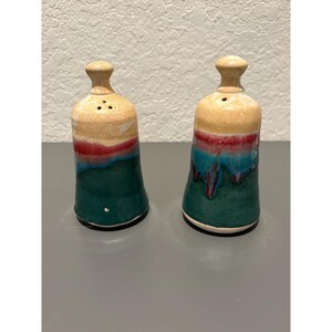 May include: A pair of ceramic salt and pepper shakers. The shakers are bell-shaped with a tan top, a red band, and a teal and green base. Each shaker has a small knob on top and three holes for dispensing.
