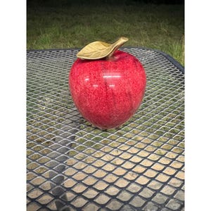 May include: A decorative apple sculpture featuring a polished, mottled red stone body with a textured surface. The apple is topped with a metallic gold-toned leaf accent that has visible vein detailing on its surface, creating a contrast in texture.