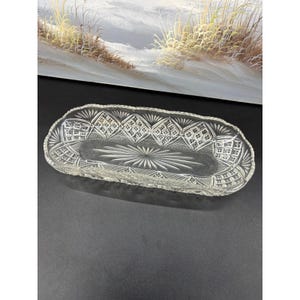 May include: A clear, rectangular glass tray featuring an intricate pressed pattern. The design includes a central starburst motif surrounded by diamond-shaped cross-hatch textures and a scalloped, serrated edge along the entire perimeter.