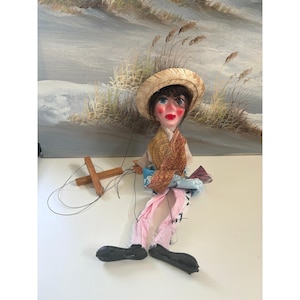 May include: A marionette puppet with dark hair, a straw hat, and painted facial features. The puppet wears a patterned brown and blue fabric outfit with light pink trousers and black shoes. It is attached to a wooden cross-shaped control bar.