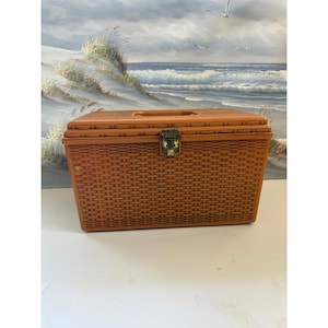 May include: A rectangular, light brown plastic storage box with a molded faux-wicker texture. The box features a hinged lid with a recessed handle and a central metal latch mechanism on the front for secure closure. The design is uniform in color.