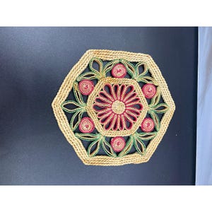 May include: A hexagonal woven trivet featuring a natural tan rope border. The centre displays a red and tan floral pattern surrounded by six small red circular accents and green leaf-shaped loops, all crafted from coiled rope material.