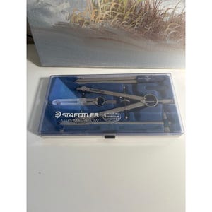 May include: A Staedtler Mars Masterbow drafting compass set housed in a clear plastic case with a blue molded interior. The set includes silver metal compasses and technical drawing tools, with the brand name and model printed on the case lid.