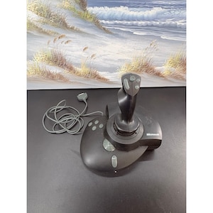 May include: A black Microsoft joystick featuring a contoured grip with multiple buttons, a circular base with additional control buttons, and a gray wired connector cable extending from the side of the device for gaming input connectivity.