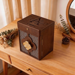 Peut inclure: Un coffret à bijoux en bois brun foncé avec un motif floral et le nom "Emma" gravé sur le couvercle. Les portes avant ont un fermoir décoratif de couleur or. La boîte est sur une table en bois.