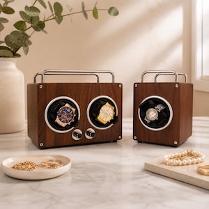 May include: Two wooden watch winders with silver handles. Each winder has a circular opening displaying a watch. One winder has two watch holders, and the other has one. The watches have gold and silver tones. A small dish with jewelry and a marble tray with a pearl necklace are in the foreground.