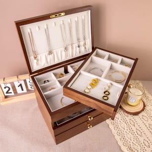 Peut inclure: Un coffret à bijoux en bois foncé avec plusieurs compartiments et tiroirs. Le coffret est ouvert, révélant des colliers, des bagues, des bracelets et d'autres bijoux. L'intérieur est doublé d'un tissu blanc doux.