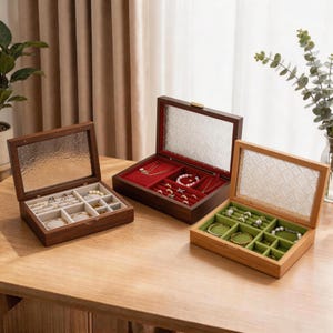Puede incluir: Tres joyeros de madera con tapas de cristal sobre una mesa de madera. Las cajas están abiertas, revelando compartimentos forrados con tela roja, verde y crema. Las cajas contienen varias joyas, incluyendo collares, anillos y pulseras.
