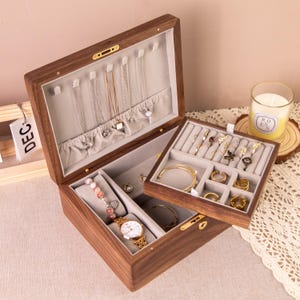 May include: A wooden jewellery box with an open lid, displaying necklaces, bracelets, rings, and a wristwatch. The box has a light grey interior and a separate tray for more jewellery. A lit candle is visible in the background.