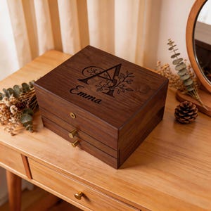 Könnte beinhalten: Eine dunkelbraune Holzschmuckschatulle mit zwei Schubladen und goldfarbenen Knöpfen. Die Oberseite der Schatulle ist mit dem Buchstaben "A", floralen Akzenten und dem Namen "Emma" graviert. Die Schatulle steht auf einem Holztisch.