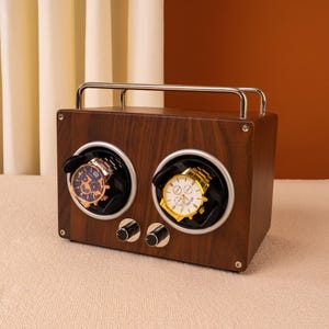 May include: A dark wooden watch winder with two compartments, each holding a wristwatch. The winder has a silver handle and two control knobs. One watch has a blue face, the other a gold face. The wood has a dark brown finish.