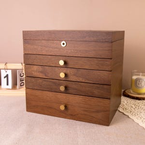 May include: A dark brown wooden jewellery box with five drawers and a top compartment. The box has gold-coloured knobs and a lock. The drawers are stacked vertically. The box is on a light-coloured surface.