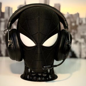 May include: A black Spider-Man themed headphone stand shaped like a mask with white eye lenses. The stand features a textured surface and a web pattern, supporting a pair of black over-ear headphones with a microphone boom arm attached to the side.