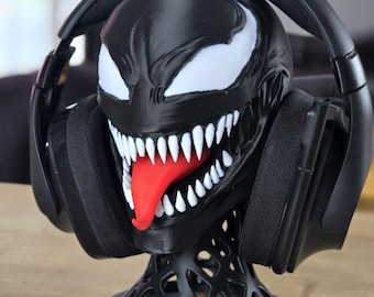 May include: A black and white Venom character head sculpture serving as a headphone stand. The figure features large white eyes, sharp white teeth, and a prominent red tongue. A pair of black over-ear headphones is resting on the head sculpture.