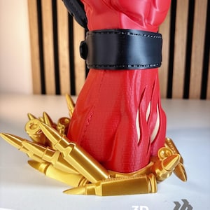 May include: A red and black fist sculpture featuring a black wrist cuff, standing on a base decorated with numerous gold-coloured bullet casings. The base includes the text 3D FACTORY BOX in white lettering at the bottom right corner of the image.
