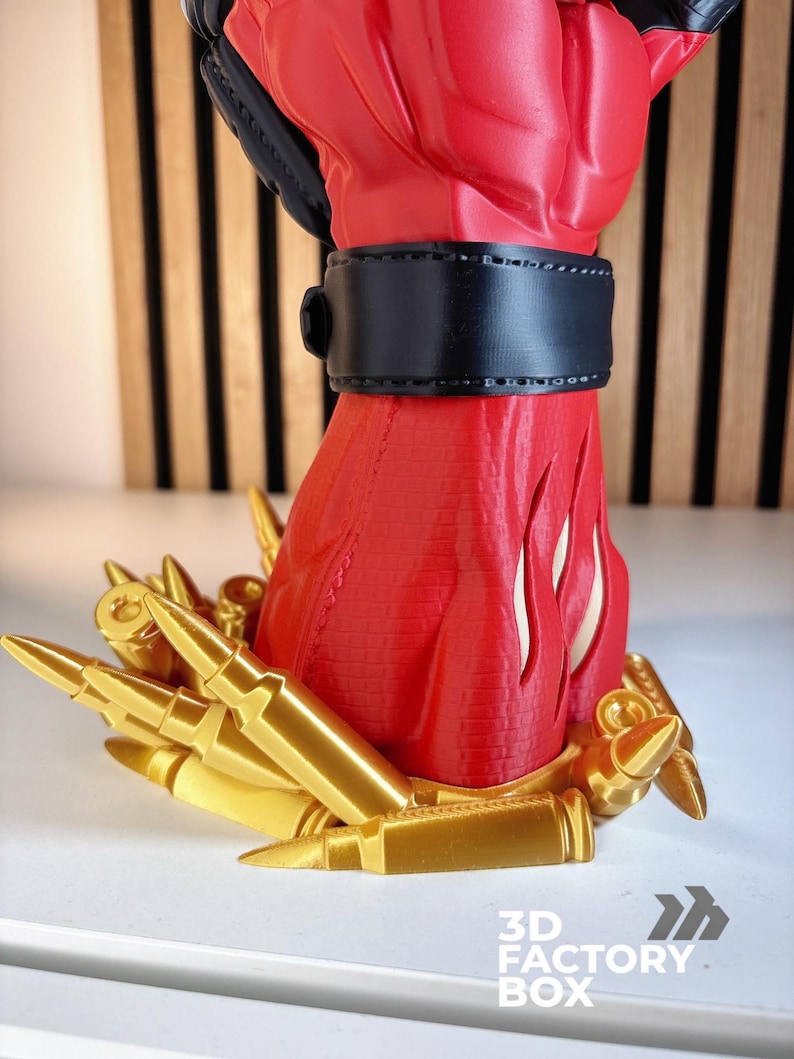 May include: A red and black fist sculpture featuring a black wrist cuff, standing on a base decorated with numerous gold-coloured bullet casings. The base includes the text 3D FACTORY BOX in white lettering at the bottom right corner of the image.