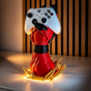 May include: A white video game controller held by a red and black glove-shaped stand. The base of the stand is surrounded by several gold-coloured bullet-shaped decorative elements, with a black band encircling the wrist area of the red glove.