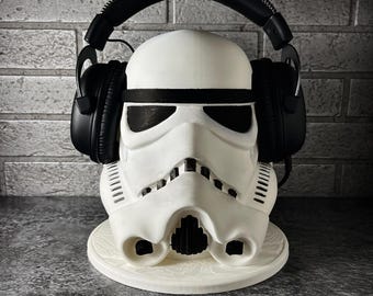 May include: A white Stormtrooper helmet display stand featuring black eye lenses and mouth details. The helmet is resting on a circular white base and is wearing a pair of black over-ear headphones with a padded headband and ear cups.