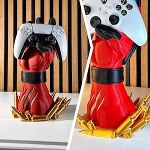 May include: A red and black sculpted hand stand designed to hold a gaming controller. The base is surrounded by gold-coloured bullet replicas. The hand features a black wrist cuff and textured details, shown holding both a white PlayStation and Xbox controller.