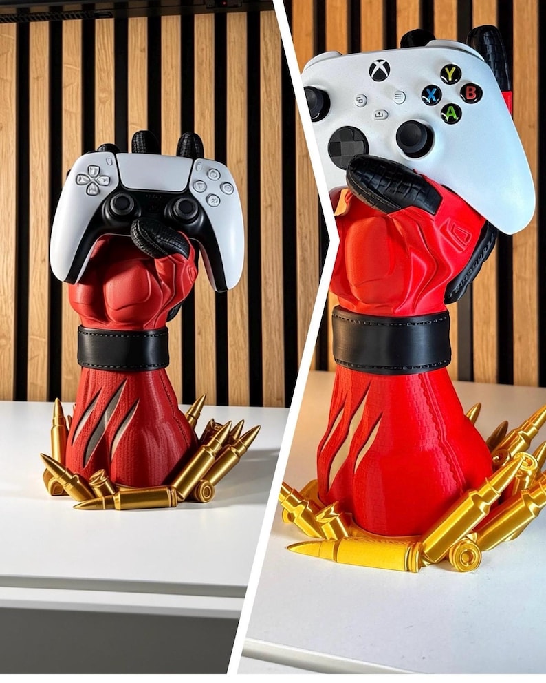 May include: A red and black sculpted hand stand designed to hold a gaming controller. The base is surrounded by gold-coloured bullet replicas. The hand features a black wrist cuff and textured details, shown holding both a white PlayStation and Xbox controller.