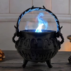 May include: A black cauldron with a three-legged base and a twisted handle. The exterior features a decorative dripping liquid design at the top and a flame pattern around the bottom. A bright blue flame rises from the center of the cauldron.
