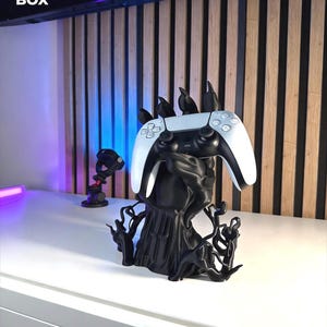 May include: A black 3D printed controller stand shaped like a clawed hand with vine-like roots at the base, holding a white and black video game controller. The text 3D FACTORY BOX is visible in the upper left corner of the image.
