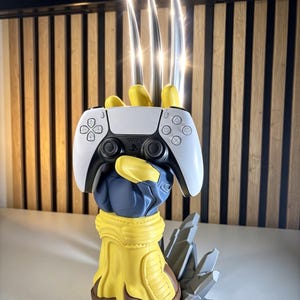 Puede incluir: Una escultura de mano enguantada en amarillo y azul con tres largas garras plateadas metálicas que se extienden hacia arriba. La mano sostiene un mando de videojuego inalámbrico blanco y negro. La base es marrón con detalles grises rocosos.