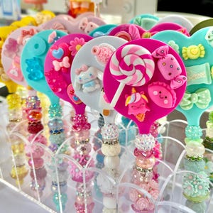Könnte beinhalten: Eine Sammlung farbenfroher, lollipopförmiger Accessoires. Jedes hat ein einzigartiges Design mit Süßigkeiten, Schleifen und Charakterverzierungen. Die Griffe sind mit Perlen und dekorativen Elementen verziert, was eine skurrile und verspielte Ästhetik schafft.