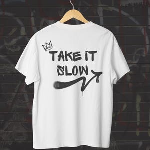 May include: White t-shirt with the phrase "TAKE IT SLOW" in black spray-paint style lettering, along with a crown graphic and an arrow. The shirt is hanging on a wooden hanger against a brick wall background.