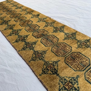 May include: A rectangular, golden-yellow runner rug with a repeating geometric pattern in shades of brown and teal. The rug features octagonal and diamond shapes, creating a symmetrical design. The rug is likely made of woven material.