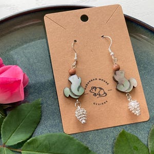 Puede incluir: Pendientes colgantes con un diseño de ardilla verde claro y dijes de piña plateados. Los pendientes se exhiben en una tarjeta marrón con el texto "Suburban Mouse Designs" y una ilustración de un ratón. Una rosa rosa está en el fondo.