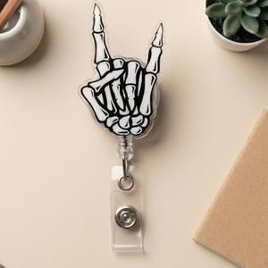 Könnte beinhalten: Ein Ausweis-Rollhalter mit einer schwarz-weißen Skeletthand, die die "Rock on"-Geste zeigt. Der Ausweis-Rollhalter hat einen transparenten Kunststoffkörper mit einem Metallclip und einem einziehbaren Kabel. Ein lustiges und kantiges Accessoire.