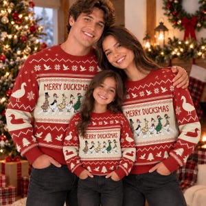 Könnte beinhalten: Ein roter Langarmpullover mit Rundhalsausschnitt, einem weißen Streifen mit dem Text MERRY DUCKSMAS und einer Grafik von sechs in einer Reihe laufenden Enten. Der Pullover zeigt wiederkehrende Muster aus weißen Enten, Schneeflocken und Tannen.