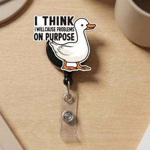 May include: A white duck badge reel with orange feet and beak, and the text "I THINK I WILL CAUSE PROBLEMS ON PURPOSE". The badge reel has a black backing, a retractable cord, and a clear plastic ID holder.