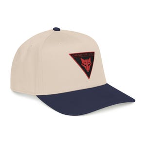 May include: A baseball cap with a cream-colored crown and a navy blue brim. The cap features a red and black embroidered patch with a wolf head design and the text "OMNIS VIRTUS". The cap has a curved brim and a button on top.