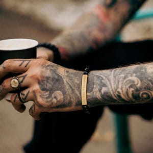 May include: A beaded bracelet featuring a rectangular gold-toned metal bar engraved with the text MY HONOR REMAINS. The bracelet is constructed with small, smooth, round black beads strung on an elastic cord, worn around the wrist.