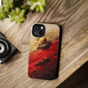 May include: A phone case featuring a science fiction landscape with a red planet, floating structures, and a large, yellow sphere. The case has a black camera lens cutout and a beige border, set against a wooden background.