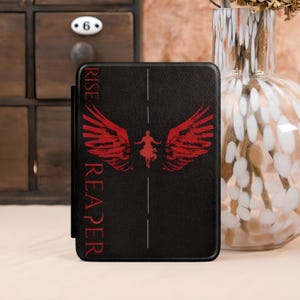 May include: Black tablet case featuring a red winged figure design with the words "RISE REAPER" in red. The case has a textured appearance and a vertical white line down the center. The design is centered on the case.