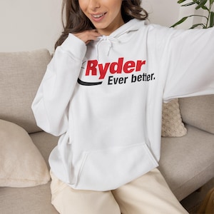 May include: A white hooded sweatshirt featuring a red and black graphic on the chest that reads Ryder Ever better. The garment includes a drawstring hood, long sleeves with ribbed cuffs, and a large front kangaroo pocket for storage.