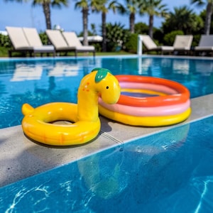 May include: A yellow inflatable pool float shaped like a giraffe with a green leaf detail, and a small, multi-colored inflatable pool. The items are placed on the edge of a pool with blue water, with lounge chairs and palm trees in the background.