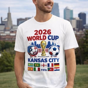 May include: White t-shirt with a graphic promoting the 2026 World Cup in Kansas City. The design includes soccer balls, a trophy, and flags. The text "2026 WORLD CUP KANSAS CITY" is prominently displayed in red and blue.
