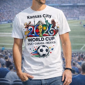 May include: White t-shirt with a colorful graphic promoting the 2026 World Cup in Kansas City, USA, Canada, and Mexico. The design includes a soccer ball, the World Cup trophy, and various national symbols.